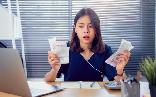 Shocked face Asian woman hand holding expense bill and calculation about debt bills monthly at the table in home office.